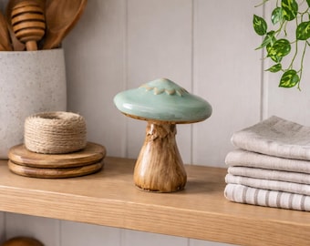 May include: A ceramic mushroom figurine with a light green cap and a brown stem. The mushroom is on a wooden shelf with other kitchen items, including wooden spoons, a honey dipper, and a stack of linen towels.