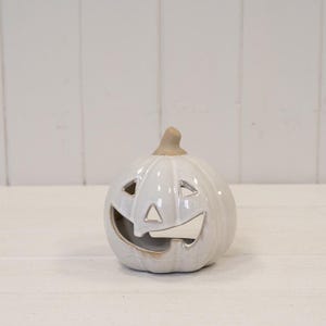 May include: A white ceramic jack-o'-lantern with a glossy finish, featuring triangular eyes, a triangular nose, and a wide, curved mouth opening. The pumpkin has a light brown, unglazed stem at the top and vertical ridges along its rounded body.