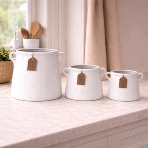May include: Three white ceramic pots of different sizes, each with small handles and brown tags. They are displayed on a countertop. The pots are suitable for kitchen or home decor.
