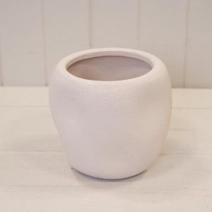 May include: A rounded, off-white ceramic planter with a textured, matte finish. The vessel features a wide, circular opening at the top and a smooth, curved body that tapers slightly towards the base, creating a simple, organic, and bulbous shape.