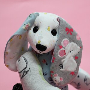 May include: A gray and blue patchwork stuffed animal with a mouse design on one ear and the word "hello!" on the other ear. The animal has a black nose and large black eyes.