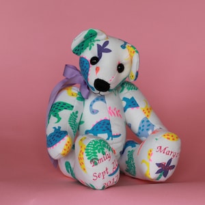 May include: A white teddy bear with a dinosaur print fabric. The bear is sitting with its legs crossed. The bear's paws are embroidered with the names "Emily" and "Marga" and the date "Sept 21, 2017".