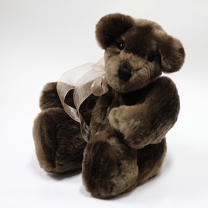 May include: A brown teddy bear with a light brown satin ribbon around its neck. The bear is sitting on a white background.