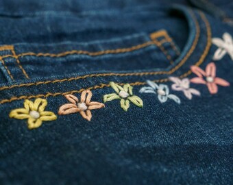 May include: Close-up of a pair of blue denim jeans with a row of embroidered flowers in pastel colors along the top of the pocket.