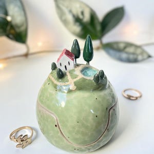 May include: A spherical ceramic trinket box with a light green textured surface. The lid features a miniature landscape with a small white house with a red roof, dark green trees, and a light blue pond. Two gold-toned rings sit nearby.
