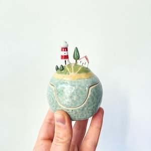 May include: A small, round, ceramic lidded jar with a light blue, textured base. The lid features a sculpted landscape with a red and white striped lighthouse, a small white house with a red roof, and a single green tree on a grassy hill.