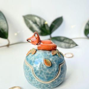 Peut inclure: Une boîte à bijoux en céramique ronde avec un vernis bleu clair texturé. Un renard sculpté orange et blanc dort sur le couvercle, entouré de petites feuilles vertes et brunes. La boîte possède un couvercle amovible et une base arrondie.