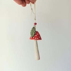 May include: A whimsical, handmade mushroom ornament. The mushroom has a red cap with white polka dots, a cream-colored stem, and a green leaf. It is suspended by a natural twine string, perfect for hanging as a decoration.