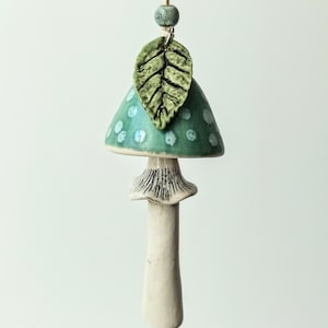 Puede incluir: Adorno de cerámica con forma de seta con un gorro turquesa con lunares blancos. Una hoja verde y una pequeña cuenta cuelgan sobre la seta, suspendidas por una cuerda de color natural. El tallo es blanquecino.