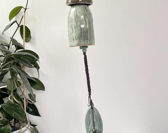 May include: A ceramic wind chime featuring a bell-shaped body in a pale green glaze with leaf imprints. It includes a dark brown dome-shaped top piece with a textured bead, a braided dark brown cord, and a leaf-shaped ceramic pendant at the base.