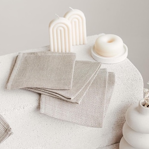 May include: A still-life arrangement featuring neutral-toned linen napkins, arch-shaped candles, and a small, round candle on a white pedestal. A stacked, white ceramic vase with dried stems is also present. The scene is minimalist and evokes a sense of calm.