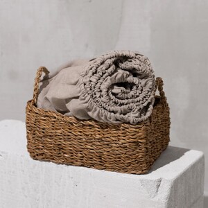 Op de afbeelding: Een rechthoekige rieten mand met een opgerolde beige lakenset. De mand, met handvatten, staat op een wit, getextureerd oppervlak. De achtergrond is een lichtgrijze muur.