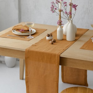 Pode incluir: Um caminho de mesa retangular de linho na cor mostarda sobre uma mesa de jantar de madeira clara. O caminho é acompanhado por jogos americanos retangulares a condizer, vasos de cerâmica branca com flores cor-de-rosa, velas e nozes.