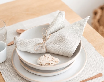 May include: A minimalist table setting with white ceramic plates, a folded linen napkin, and copper-colored forks. The napkin is tied with twine and rests on a stack of plates. A small, white cup and a glass are also present.