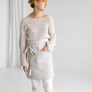 Puede incluir: Una mujer lleva un delantal de lino beige con un bolsillo sobre un suéter de crochet blanco y pantalones blancos. Está de pie en una habitación blanca con una cortina blanca en el fondo.