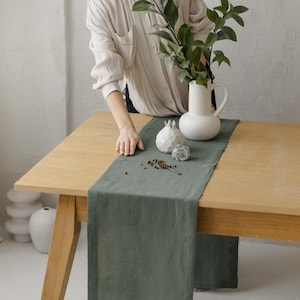 Pode incluir: Um caminho de mesa longo e retangular de linho em verde sálvia, colocado sobre uma mesa de madeira clara. O caminho apresenta uma textura lisa e mate com bordas bainhadas limpas, oferecendo uma base de cor sólida para decoração e acessórios.