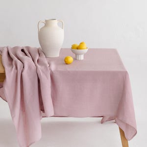 Può includere: Una tovaglia in lino rosa antico su un tavolo di legno, con un vaso color crema e una piccola ciotola bianca di limoni. L'apparecchiatura è semplice ed elegante, con un'estetica minimalista.