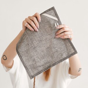 May include: A square, gray and black checkered textile with a white ribbon and tag. The textile is held up by a person. The textile has a dark border and a small tag with a leaf design.