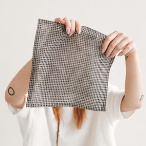 May include: A square, checkered fabric in shades of gray and black is held up in front of a person. The fabric has a small, repeating pattern. The person is wearing a white t-shirt.