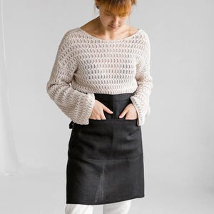 May include: A black apron with two front pockets. The apron is worn over white pants and a cream-colored sweater. The sweater has a loose knit design. The person is standing in a white room.