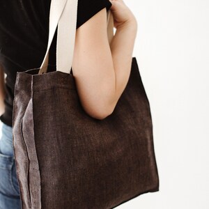 May include: A brown canvas tote bag with cream colored straps. The bag is being held by a person wearing a black shirt and blue jeans.