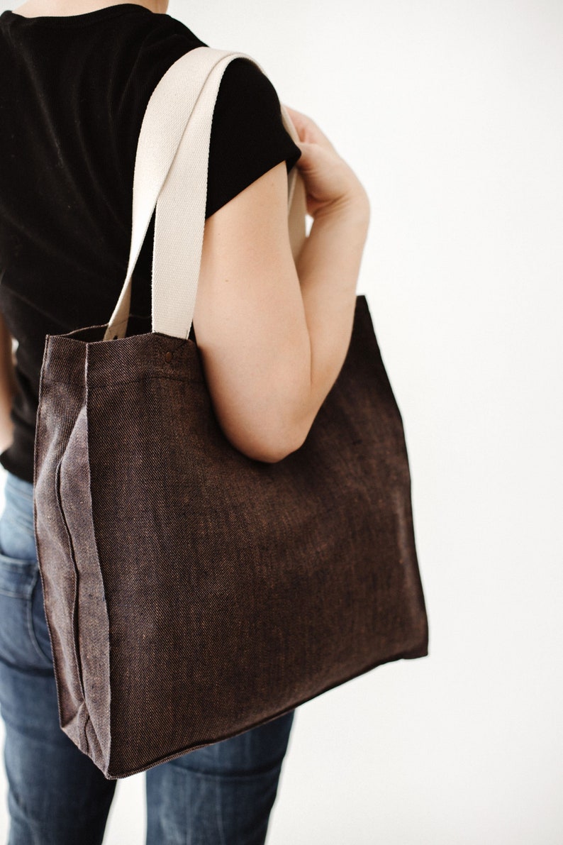 May include: A brown canvas tote bag with cream colored straps. The bag is being held by a person wearing a black shirt and blue jeans.