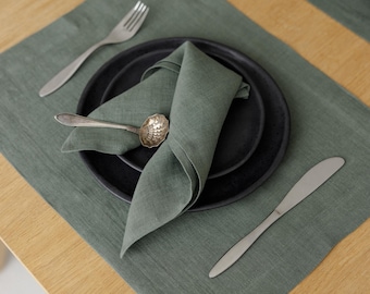May include: A sage green linen placemat and matching folded napkin set on a wooden table. The setting includes two matte black ceramic plates, a silver fork, a silver knife, and a small decorative silver spoon with a textured bowl design.