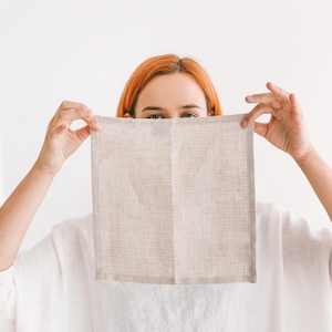 May include: A person holds a beige linen tea towel in front of their face. The towel is folded in half and has a subtle texture.