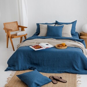 Pode incluir: Uma cena de quarto com uma cama com roupa de cama azul, almofadas bege e um cobertor bege. Uma cadeira de madeira, um tapete pequeno e uma bandeja com bebidas e lanches também são visíveis. O quarto tem uma estética minimalista e acolhedora.