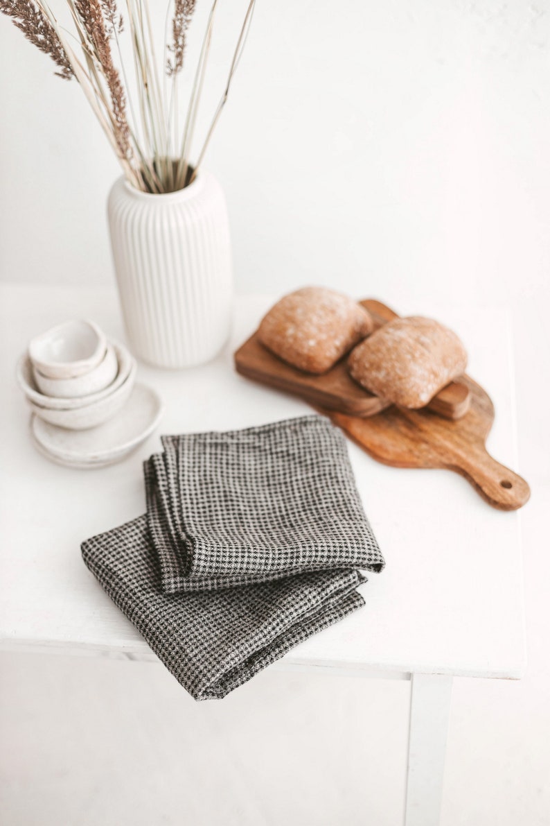 May include: A still-life arrangement featuring a white vase with dried plants, small white bowls, and two folded gray and black checkered napkins. Two loaves of bread rest on a wooden cutting board. The scene is set on a white surface.