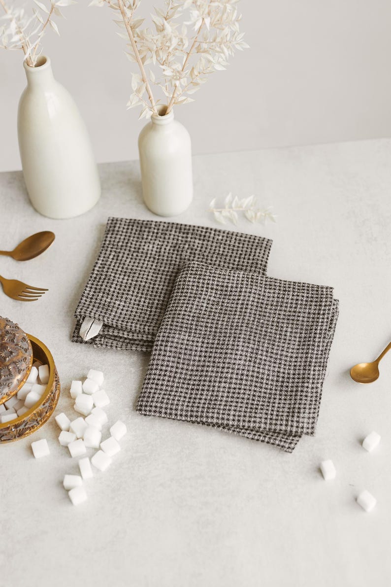 May include: Two folded gray and white checkered napkins are displayed on a white surface. The napkins are next to a small white vase with dried flowers, a gold spoon, and a decorative box with sugar cubes.