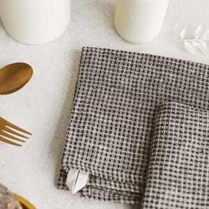 May include: A stack of two folded black and white checkered napkins with a small white tag. The napkins are on a light gray surface, with a gold fork and spoon, and white ceramic vases in the background.