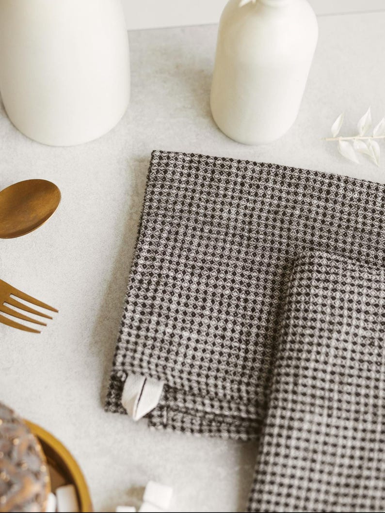 May include: A stack of two folded black and white checkered napkins with a small white tag. The napkins are on a light gray surface, with a gold fork and spoon, and white ceramic vases in the background.