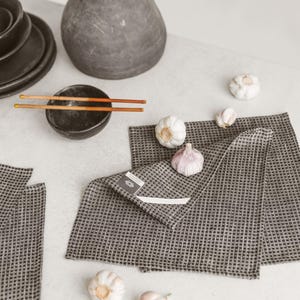 May include: A still-life arrangement featuring a dark gray vase with dried stems, a small black bowl with chopsticks, and several heads of garlic. Two gray checkered kitchen towels are folded on a white surface.