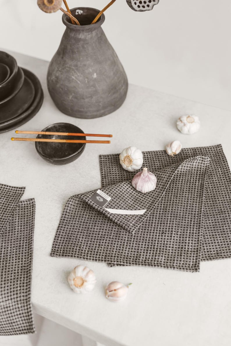 May include: A still-life arrangement featuring a dark gray vase with dried stems, a small black bowl with chopsticks, and several heads of garlic. Two gray checkered kitchen towels are folded on a white surface.
