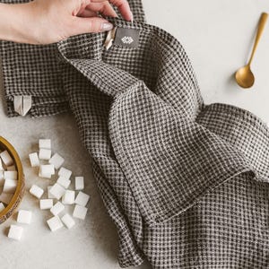 May include: A folded brown and white checkered kitchen towel with a tag, next to a stack of similar towels. A gold spoon and a small wooden bowl with sugar cubes are also visible.