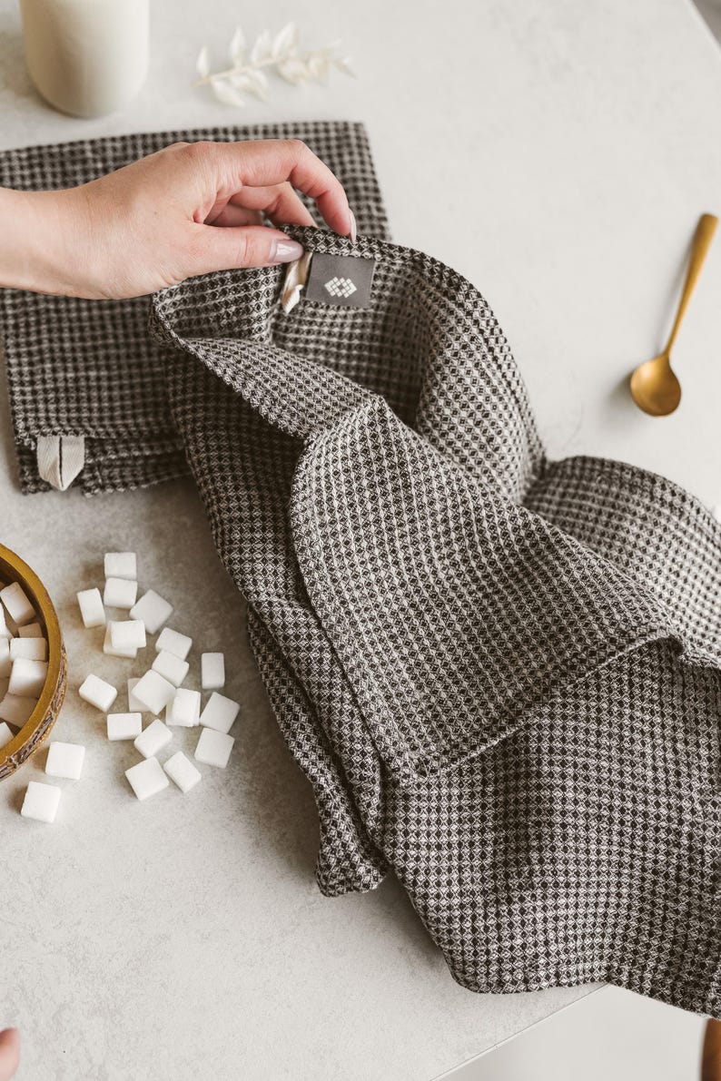 May include: A folded brown and white checkered kitchen towel with a tag, next to a stack of similar towels. A gold spoon and a small wooden bowl with sugar cubes are also visible.