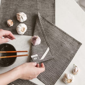 May include: A top-down shot shows a set of gray checkered kitchen towels with several heads of garlic. A black bowl with chopsticks is in the frame. The towels are folded and arranged on a white surface.