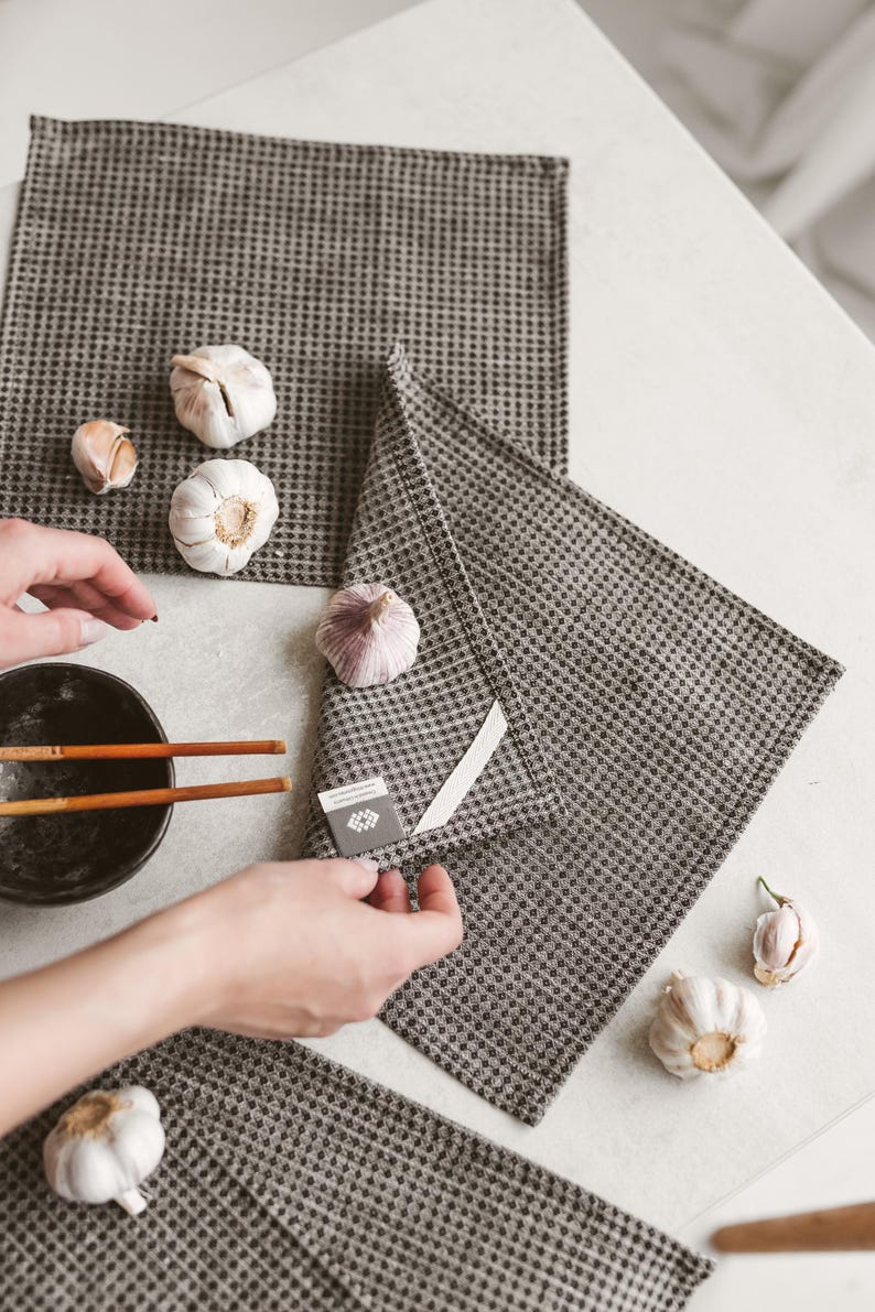 May include: A top-down shot shows a set of gray checkered kitchen towels with several heads of garlic. A black bowl with chopsticks is in the frame. The towels are folded and arranged on a white surface.