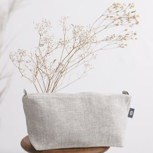 A large white linen makeup bag with a zipper decorated in a herringbone pattern on a wooden stool.