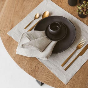 Könnte beinhalten: Tischarrangement mit hellgrauem Leinen-Tischset, schwarzen Keramikplatten und goldfarbenem Besteck. Das Arrangement umfasst eine gefaltete Serviette, eine kleine Schüssel und zwei Löffel. Dekorative schwarze Vasen und eine kleine Pflanze befinden sich im Hintergrund.