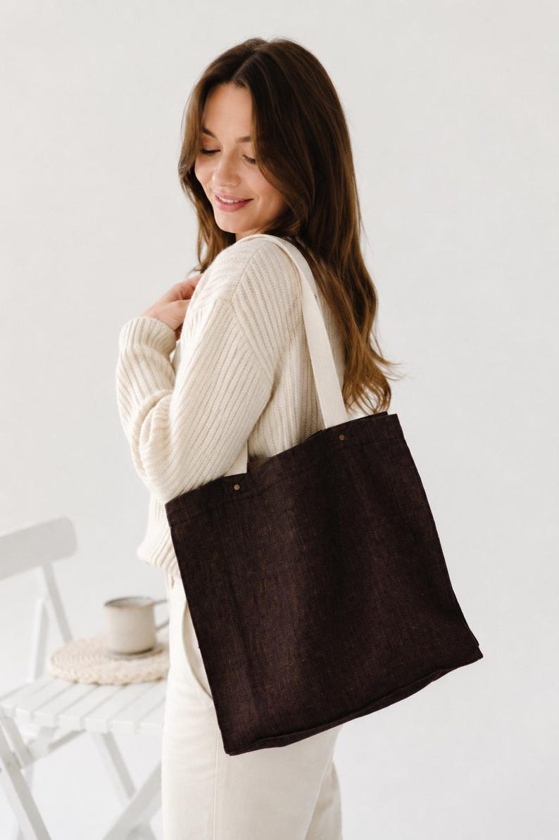 May include: A dark brown tote bag with a visible woven texture and two off-white fabric shoulder straps. The bag features a rectangular shape with reinforced stitching at the top corners where the straps are attached with small metallic rivets.