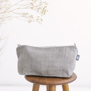 A large light grey linen toiletry bag in herringbone pattern with a zipper on a stool