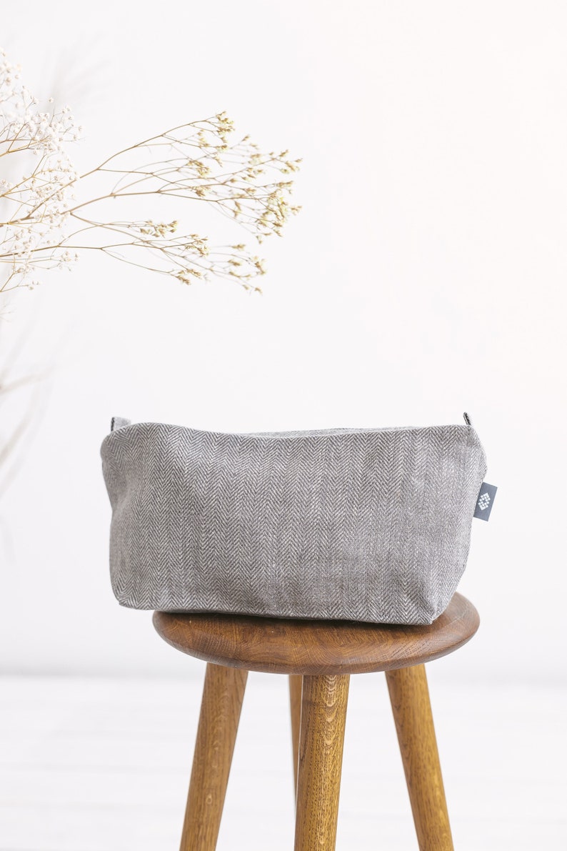 A large light grey linen toiletry bag in herringbone pattern with a zipper on a stool