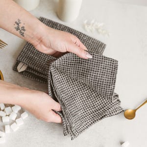 May include: A stack of black and white checkered napkins. The napkins are being held by a person. A gold spoon and white vases are in the background. Sugar cubes are scattered on the surface.
