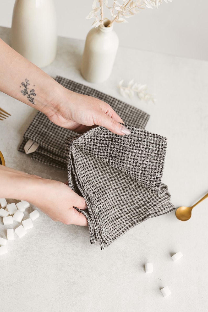 May include: A stack of black and white checkered napkins. The napkins are being held by a person. A gold spoon and white vases are in the background. Sugar cubes are scattered on the surface.