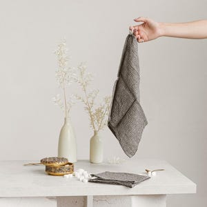 May include: A gray and white checkered linen napkin is held up by a hand, with a stack of napkins on a white surface. A small, decorative box and two white vases with dried flowers are also on the surface.