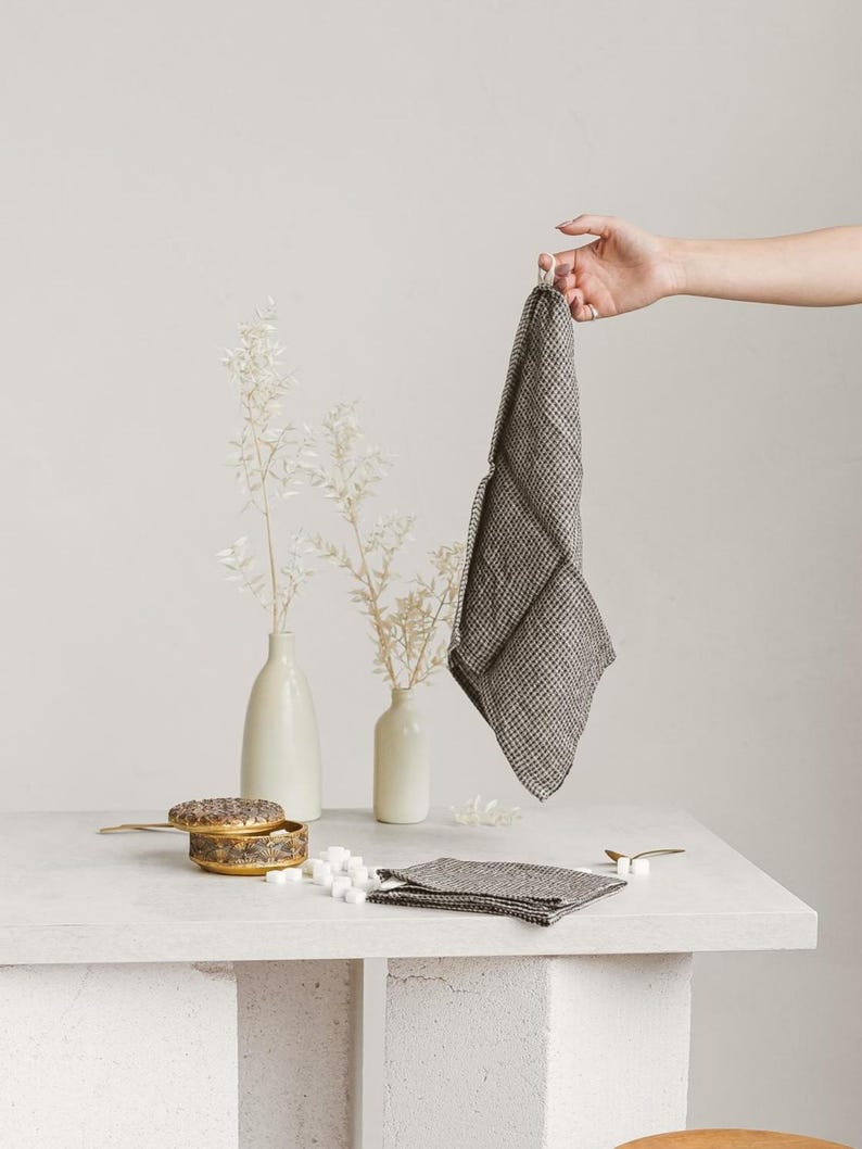 May include: A gray and white checkered linen napkin is held up by a hand, with a stack of napkins on a white surface. A small, decorative box and two white vases with dried flowers are also on the surface.