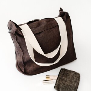 May include: A brown canvas tote bag with white straps. The bag is lying on a white surface with a small brown and black patterned pouch and two gold and silver tubes.