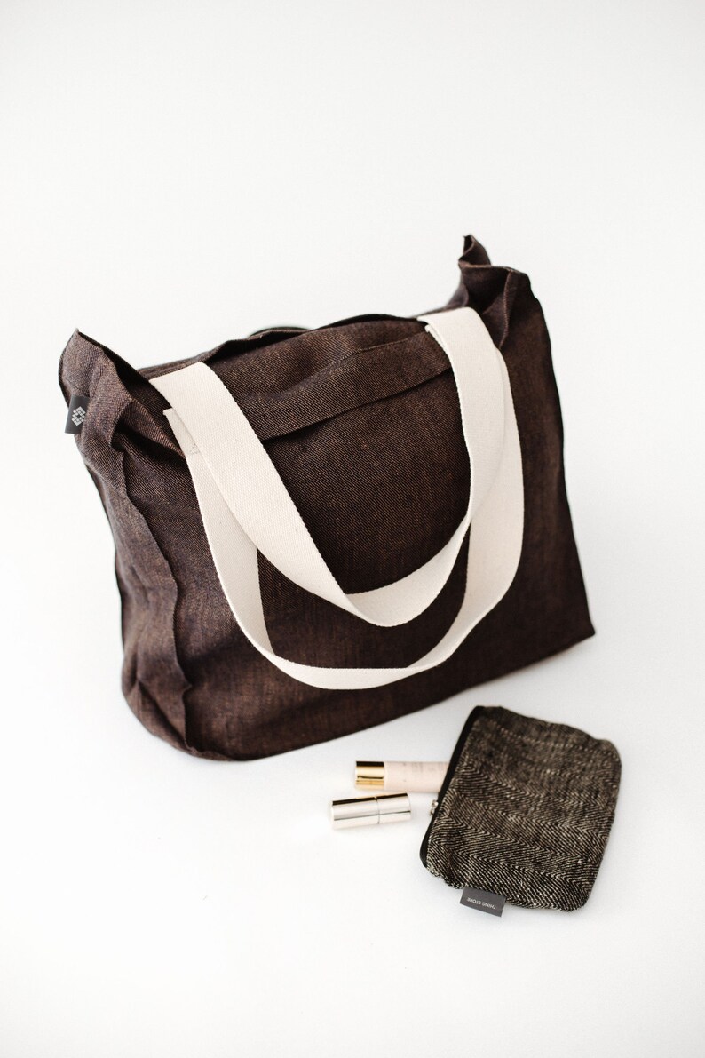 May include: A brown canvas tote bag with white straps. The bag is lying on a white surface with a small brown and black patterned pouch and two gold and silver tubes.